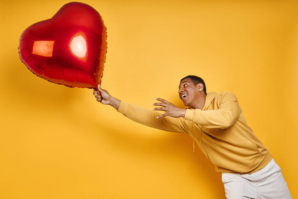 Playful mixed race man catching big heart shape balloon while standing against yellow background - Foto, Imagen