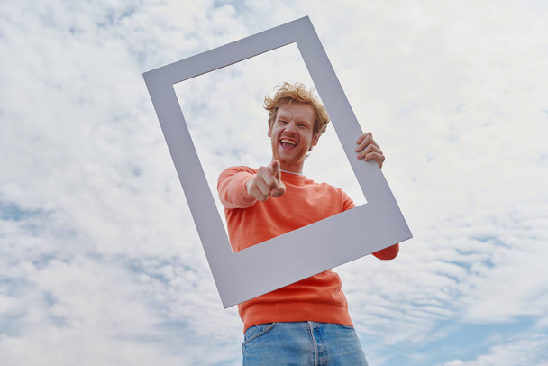 Playful young redhead man looking through a picture frame and gesturing while standing outdoors - Photo, Image