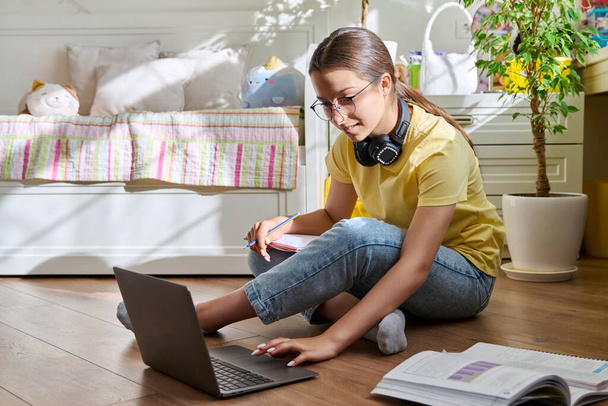 Teenage girl with glasses studying at home using a laptop. Female with headphones and textbooks sitting on the floor in the room. Education, adolescence, high school concept - Zdjęcie, obraz