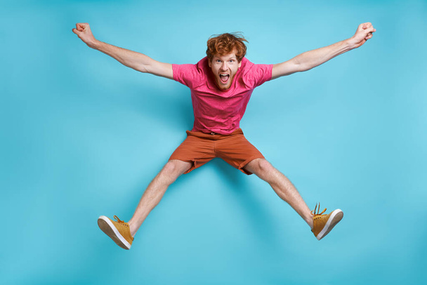 Cheerful young redhead man jumping against blue background - Fotografie, Obrázek