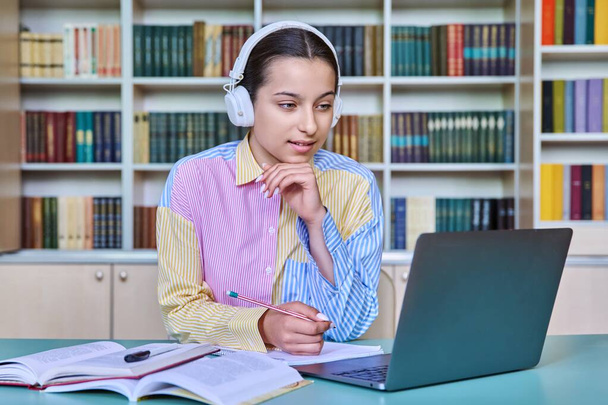 Teenage female student in headphones sitting in school library using laptop for video call conference chat looking at webcam. Online lesson, course, web seminar. Technology, e-learning, adolescence - Foto, afbeelding