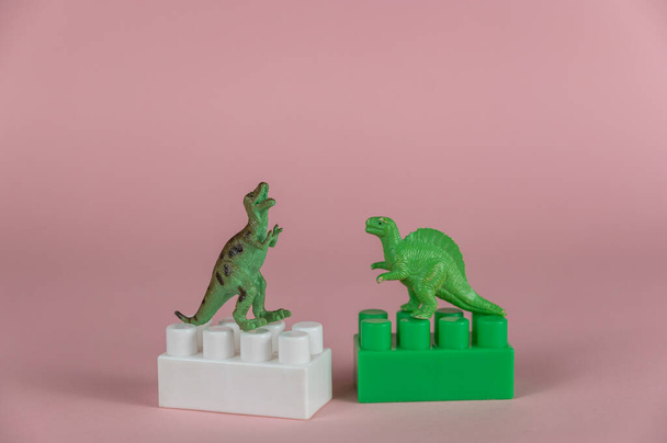 Dinosaurios en miniatura parados en dos bloques de constructor infantil. Dos pequeñas miniaturas verdes de dinosaurios depredadores de pie sobre sus patas traseras. Fondo rosa. Enfoque selectivo. - Foto, Imagen