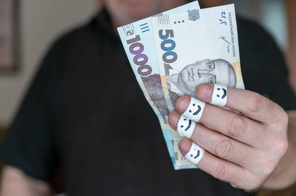 Um homem adulto tem dinheiro na mão. Algumas notas de hryvnia ucranianas. As primeiras falanges de seus dedos são envoltas em fita branca. Rostos sorridentes são pintados nas ligaduras. - Foto, Imagem