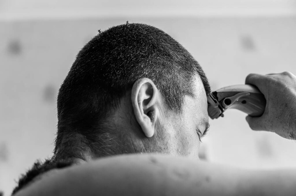 A middle-aged man cuts his hair. The person cuts the hair on his head with an electric hair clipper. Short brown hair. Inside a living room. Selective focus. Black and white photo. - Photo, Image