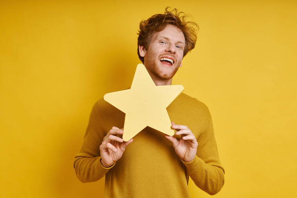 Handsome redhead man holding star shape banner and smiling against yellow background - Foto, Imagem