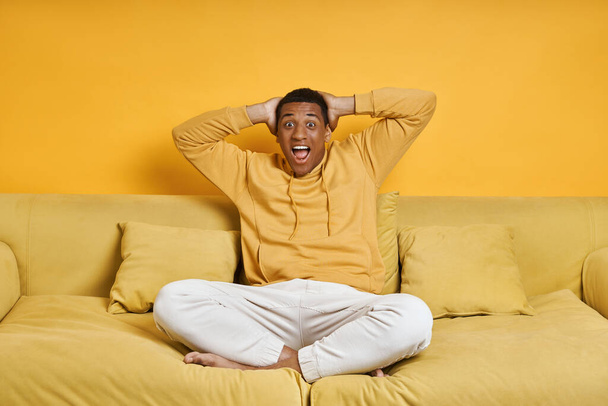 Surprised young man holding hands behind head while sitting on the couch with yellow background - Fotoğraf, Görsel
