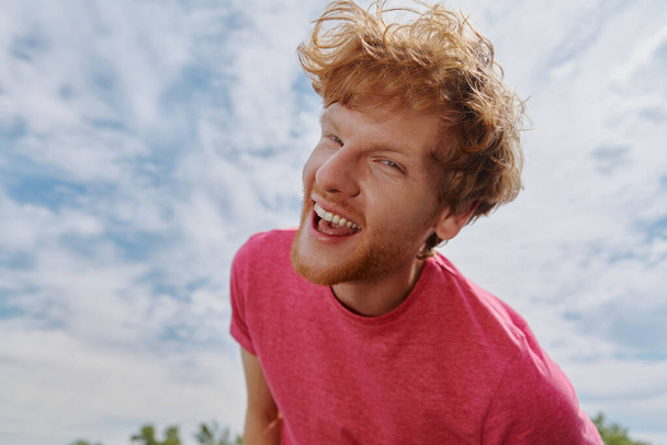 Красивый молодой рыжий мужчина смотрит в камеру и улыбается на фоне неба - Фото, изображение