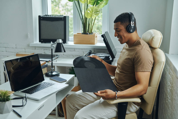 Handsome mixed race man in headphones holding vinyl record while sitting at his working place - Fotó, kép