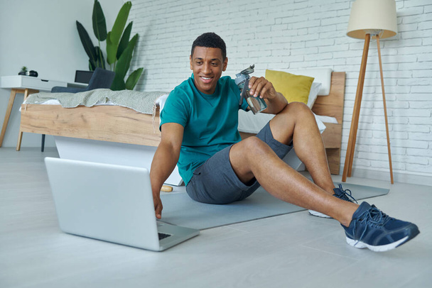 Happy young multiracial man using laptop and enjoying fresh water after training at home - Фото, изображение
