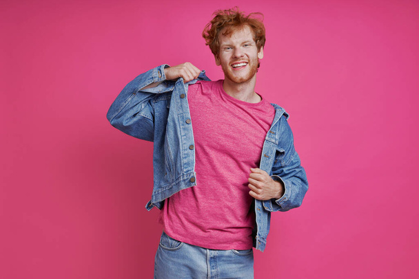 Happy redhead man in denim clothing looking at camera while standing against pink background - Photo, image