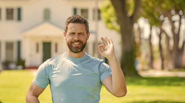 szczęśliwy Dojrzałe człowiek pośrednik stoi na zewnątrz w domu pokazując ok gest, kupując dom. - Zdjęcie, obraz