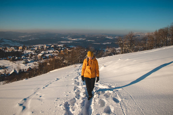 Ταξιδιώτης με ένα ευχάριστο χαμόγελο σε ένα διακριτικό κίτρινο σακάκι στέκεται στην κορυφή ενός λόφου στα βουνά της Πολωνίας Beskydy το ηλιοβασίλεμα, απολαμβάνοντας τη στιγμή. Πραγματικοί άνθρωποι σε χειμερινό πάγο περιβάλλον. - Φωτογραφία, εικόνα