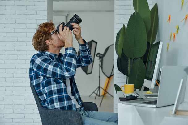 Hombre seguro examinando la cámara mientras está sentado en su lugar de trabajo en el estudio - Foto, Imagen