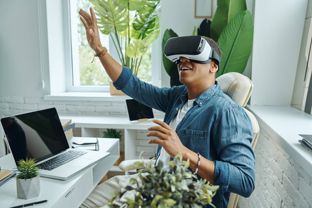 Joyful mixed race man wearing virtual reality glasses and gesturing while sitting at the desk - Фото, изображение