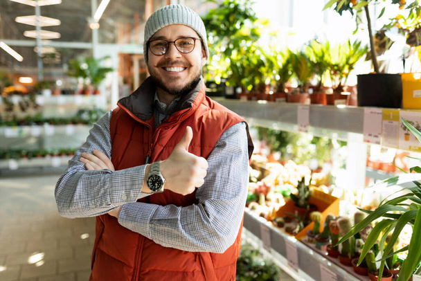 un asistente de ventas en una tienda de productos de jardinería mira a la cámara con una sonrisa y muestra un símbolo similar. - Foto, Imagen