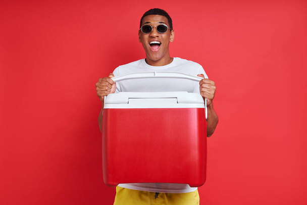 Szczęśliwy mieszane wyścig człowiek niosąc chłodniejsze pudełko stojąc na czerwonym tle - Zdjęcie, obraz