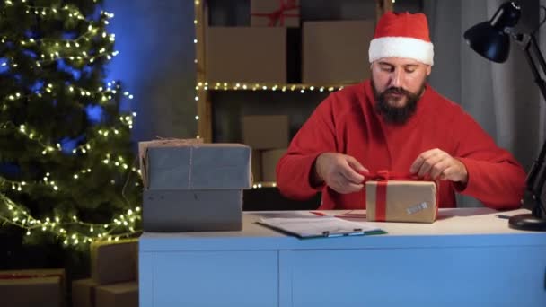 Dropshipping owner male Hindu Santa Claus works at night in his home office, knitting a big red bow on a box before shipping to a customer during the Christmas sale. Online store and small business. - Footage, Video