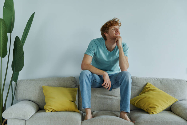 Thoughtful redhead man holding smart phone while relaxing on the couch at home - Foto, immagini