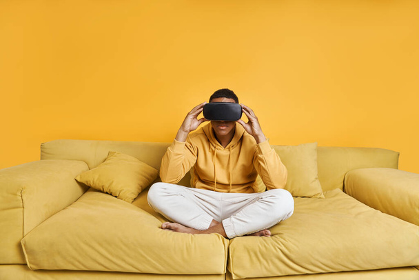 Με αυτοπεποίθηση νεαρός άνδρας σε γυαλιά εικονικής πραγματικότητας χαλαρώνοντας στον καναπέ με κίτρινο φόντο - Φωτογραφία, εικόνα
