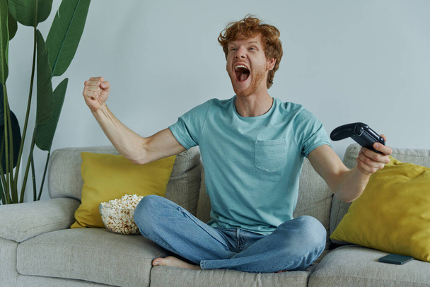 Excited redhead man playing video games and gesturing while sitting on the couch at home - Foto, Bild