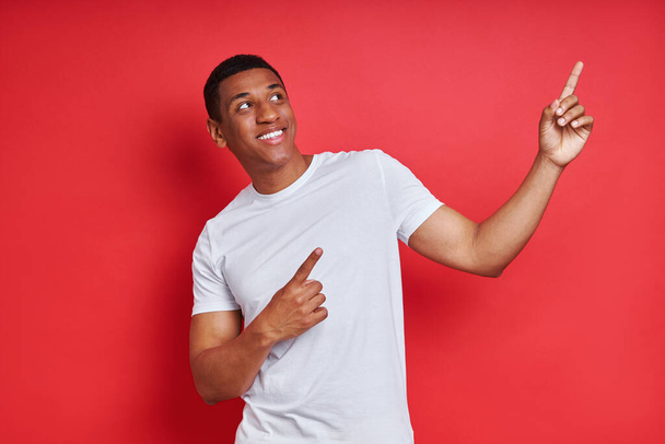 Cheerful mixed race man pointing away while standing against red background - Фото, зображення