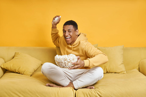 Playful mixed race man throwing popcorn while relaxing on the couch with yellow background - Foto, imagen