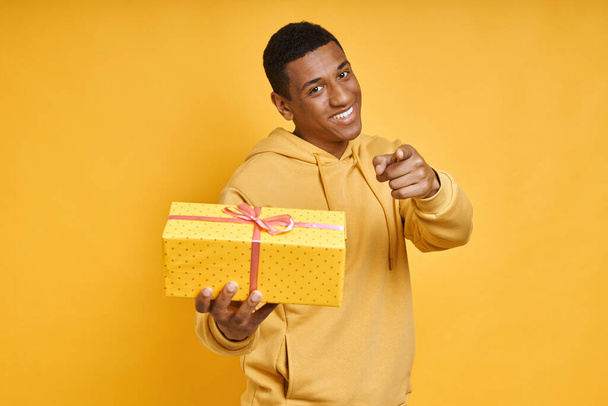 Handsome mixed race man giving a gift box and pointing while standing against yellow background - 写真・画像