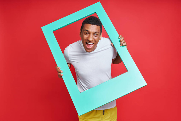 Playful mixed race man looking through a picture frame while standing against red background - Фото, изображение