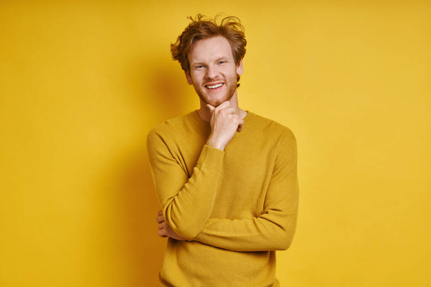 Cheerful redhead man holding hand on chin while standing against yellow background - Foto, Imagen