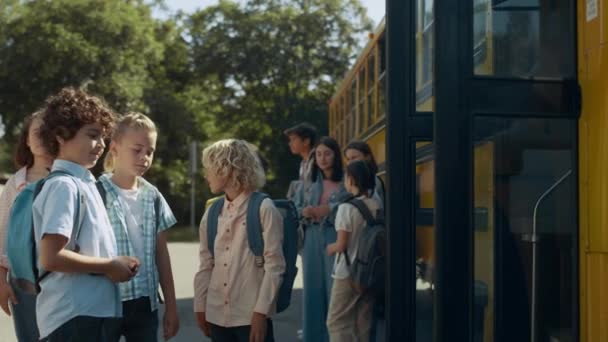 Diversos estudiantes de primaria de pie juntos en el clásico autobús escolar amarillo. Felices colegiales hablando cerca del vehículo de la puerta abierta. Alumnos aburridos esperando el día soleado del autobús. Concepto de estudio. - Imágenes, Vídeo