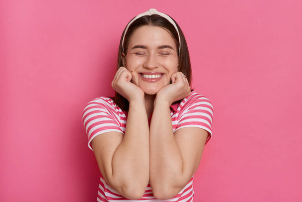 Image of smiling happy kind woman wearing striped T-shirt and hair band, keeping eyes closed, keeps hands under chin, feels happy, posing isolated over pink background. - Foto, imagen