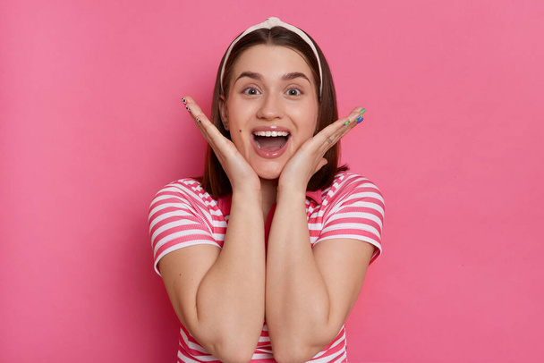 Portrait of charming happy excited woman wearing striped T-shirt and hair band posing isolated over pink background, keeps hands near face, sees something pleasant surprise. - Fotoğraf, Görsel
