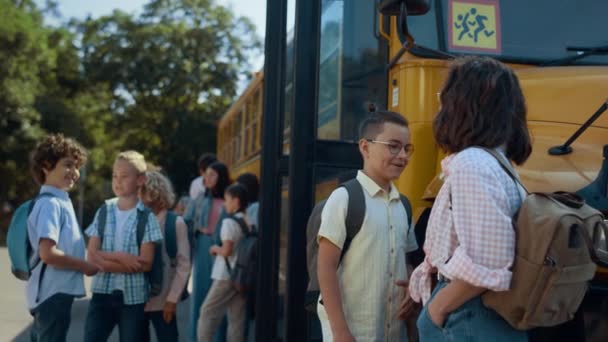 Estudiantes de primaria esperando abordar el autobús escolar. Diversos alumnos de pie hablando en el transbordador amarillo. Alegre escolares sonriendo charlando juntos. Adolescentes que se comunican después de clases al aire libre - Metraje, vídeo