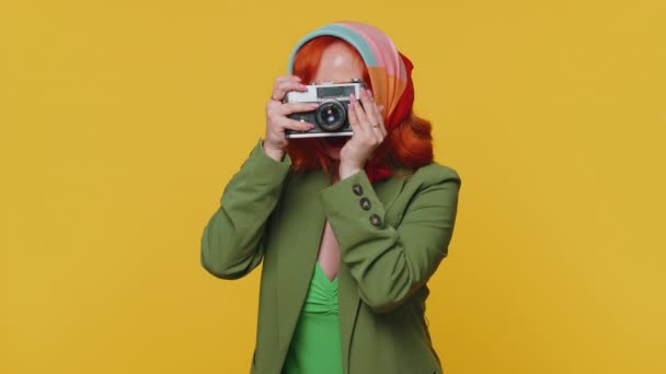 Yeşil ceketli ve elbiseli kızıl saçlı genç kadın fotoğrafçı retro kamerada fotoğraf çekiyor ve gülümsüyor. Seyahat, yaz tatili. İçerideki kızıl kız sarı stüdyo arka planında izole. - Video, Çekim