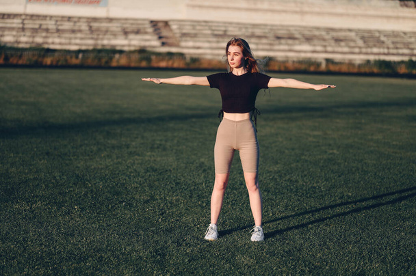 Girl in Black Top está haciendo ejercicios con los brazos a un lado. Mujer de altura completa de pie en el estadio en Green Lawn. Calentarse antes de correr. - Foto, Imagen
