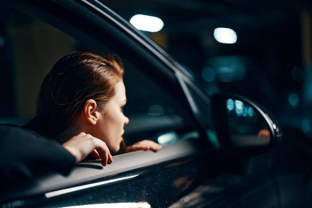 близьке горизонтальне фото з боку, вночі, жінки, що сидить у чорному автомобілі і дивиться з вікна, дивлячись у дзеркало вигляду збоку. Високоякісна фотографія - Фото, зображення