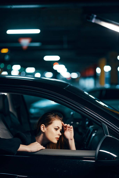 foto vertical desde un lado, por la noche, de una mujer sentada en un coche negro y mirando por la ventana mirando al espejo lateral con la mano cerca de la cara. Foto de alta calidad - Foto, Imagen