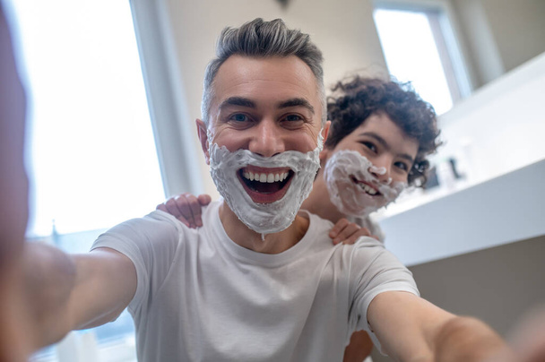 Funny shaving. Dad and son looking funny with shaving foam on their faces - Foto, afbeelding