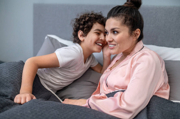 Happy morning. A cute curly-haired boy sitting in bed with his mom - Foto, afbeelding