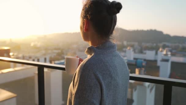 Vrouw begint haar dag met een kopje thee of koffie op het balkon bij dageraad, slow motion. Stadslandschap op de achtergrond. Moderne stedelijke levensstijl - Video