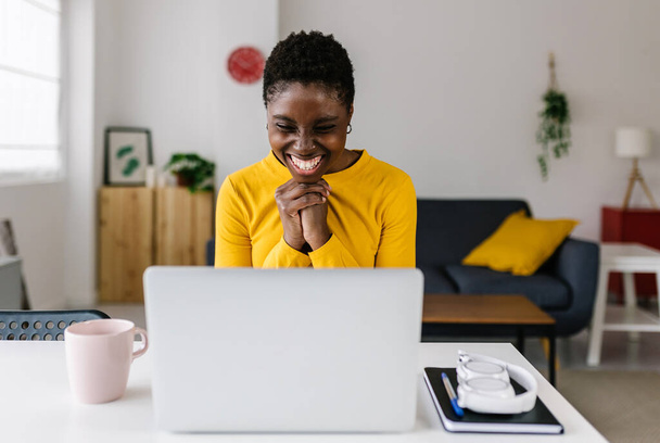 Mujer africana joven emocionada celebrando el éxito mirando la pantalla del ordenador portátil - Mujer milenaria leyendo buenas noticias en la computadora mientras está sentada en el lugar de trabajo - Concepto de victoria y celebración - Foto, imagen