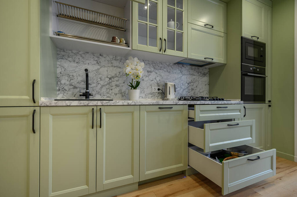 Μοντέρνο μοντέρνο ανοιχτό πράσινο πολυτελή κουζίνα με τραπεζαρία και σκούρο πράσινο καρέκλες, μερικά συρτάρια είναι ανασυρθεί - Φωτογραφία, εικόνα