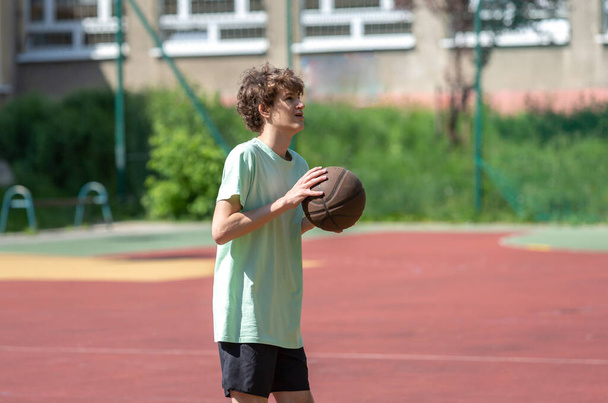 Teenager hraje basketbal s úsměvem a vesele. Fit boy s basketbalovým míčem venku. Kid dribling the ball at the court - Fotografie, Obrázek