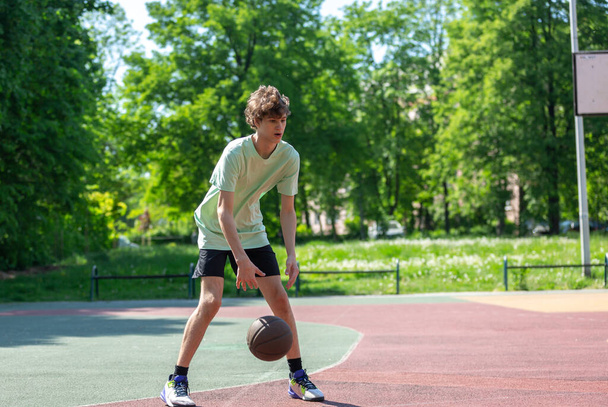 Basketbol oynayan genç, gülümseyen, neşeli. Basketbol topu olan zinde bir çocuk. Sahada top süren çocuk. - Fotoğraf, Görsel