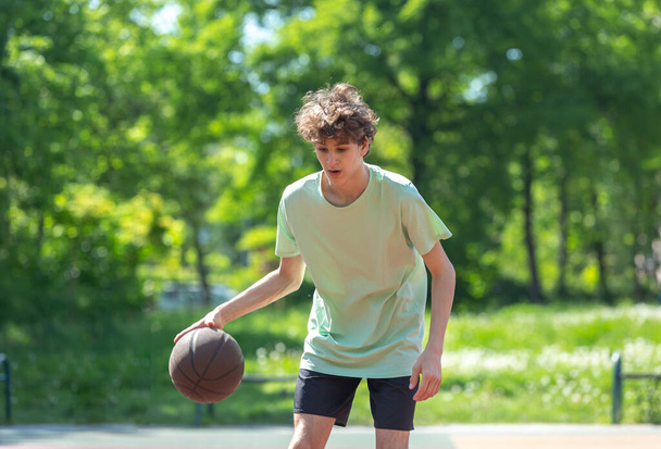 Teenager playing basketball smiling, cheerful. Fit boy with basketball ball outdoors. Kid dribbling the ball at the court - Foto, Imagen
