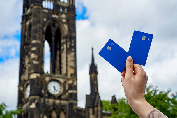 Молодые женщины держат голубые банковские карточки на фоне неба и старого здания часовни. Банковские карты с чипом и бесконтактной функцией оплаты. Платежи. - Фото, изображение