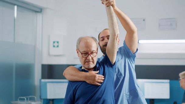 Medische assistent het verhogen van de armen van oudere persoon te rekken spieren en de behandeling van lichamelijk letsel. Osteopathie specialist doet mobiliteitsoefening met senior man bij fysiotherapie afspraak. - Foto, afbeelding
