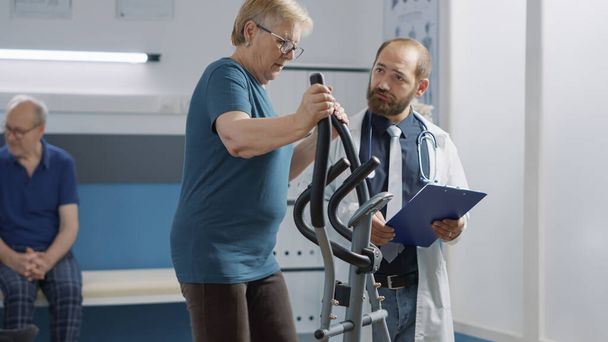 Gyakorló asszisztál idős nő rehabilitációs fizioterápiás eljárás, gyakorló álló elektromos kerékpár. Az orvos segít a betegnek a sérülés gyógyításában és a mobilitás növelésében. - Fotó, kép