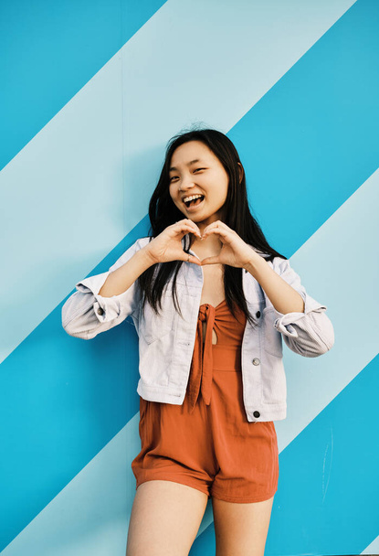 Happy young Asian woman makes a heart shape with her hands while standing outdoors against a striped wall. Love and emotions concept. - Photo, Image