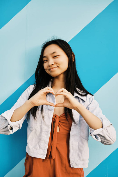 Portret młodej Azjatki patrzącej w kamerę i tworzącej serce dłońmi, stojącej przy ścianie w paski. Koncepcja miłości i emocji. - Zdjęcie, obraz